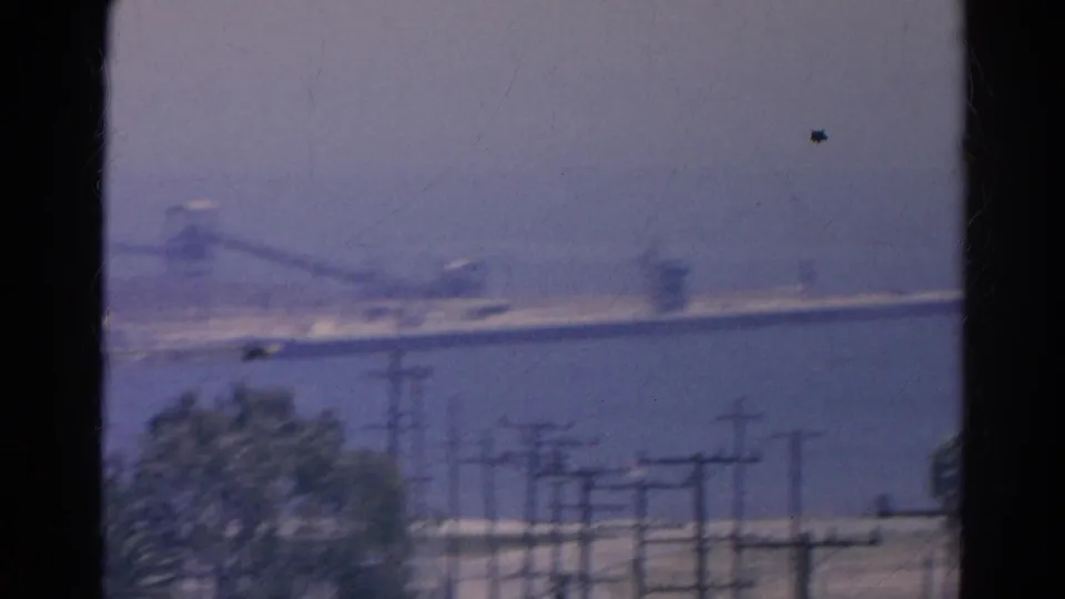 1966 home-movie frame with utility poles and power lines in front of water and a distant bridge.