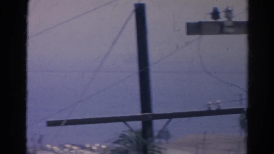 Blue-tinted 1966 home-movie frame showing a telephone crossarm, vertical pole, overhead wires, and palm fronds near a distant horizon.