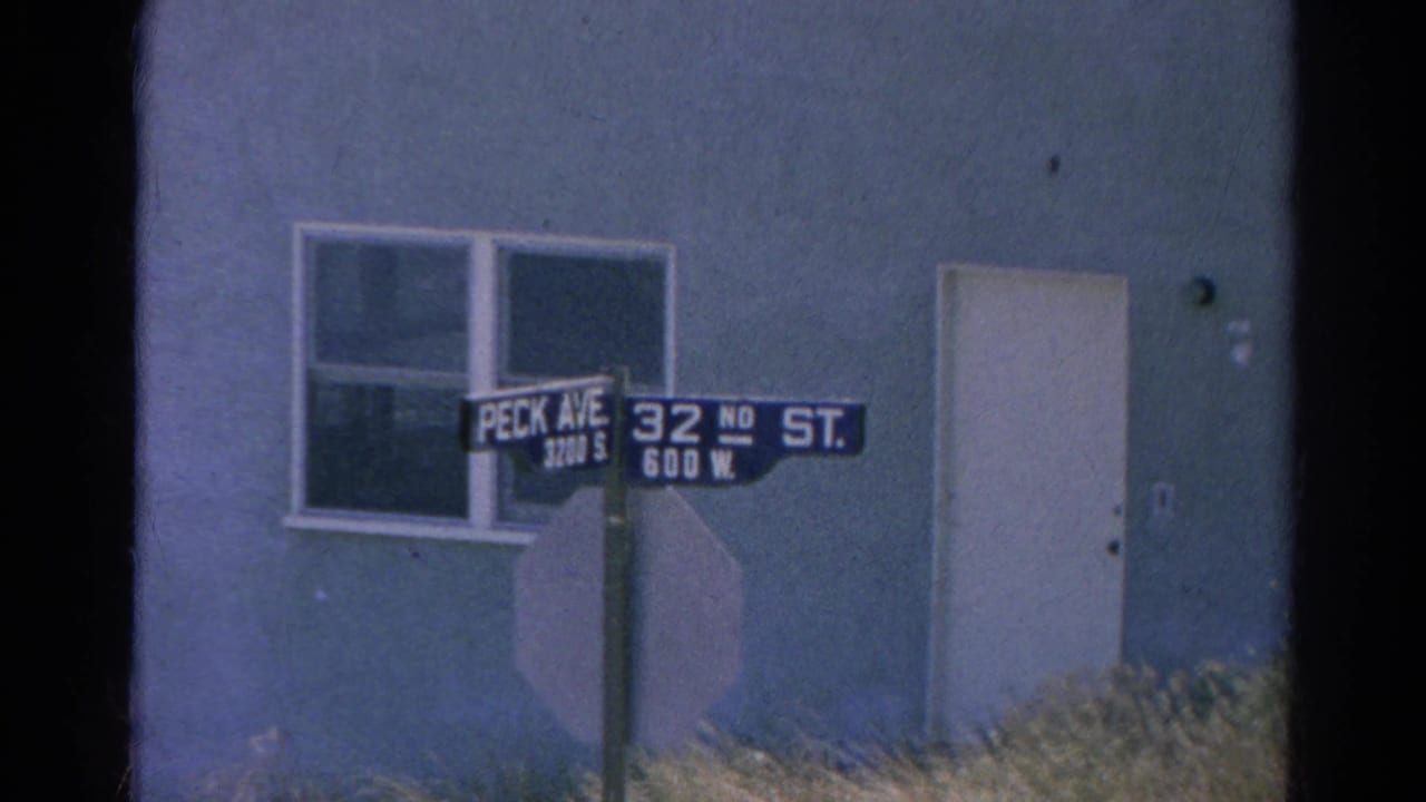 Street signs for Peck Ave and 32nd St stand before a plain building wall with a window and door.