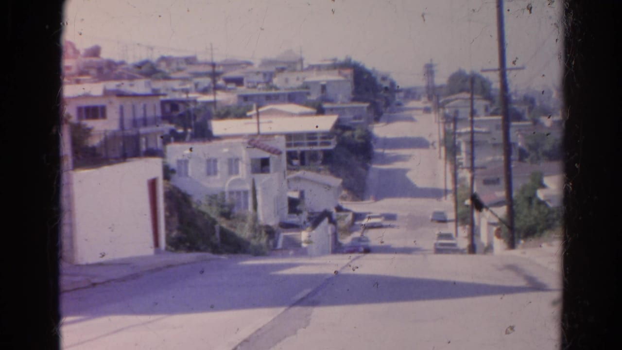 Steep residential street with stucco houses, parked cars, utility poles, and overhead wires.