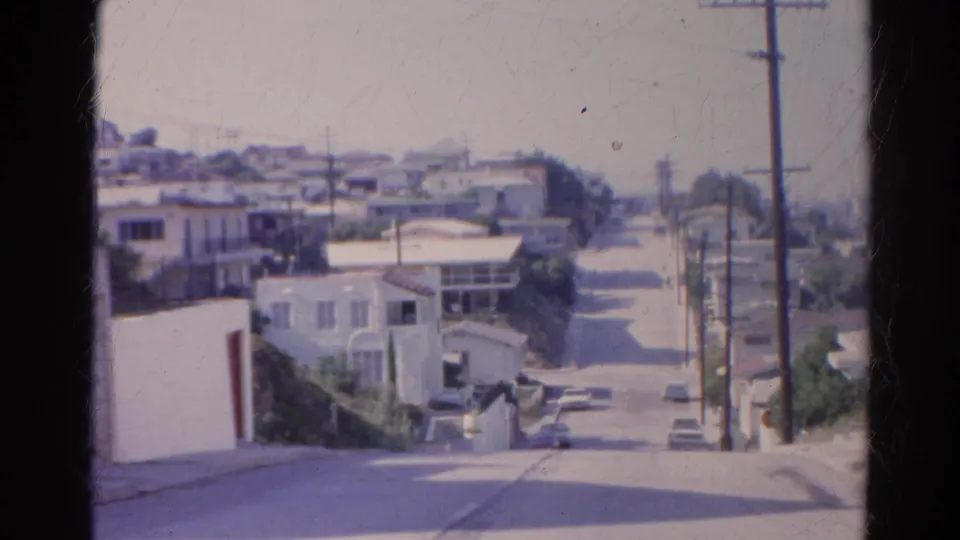 1966 home-movie frame of a Silver Lake hillside street with stucco houses, garages, parked cars, poles, and overhead wires.