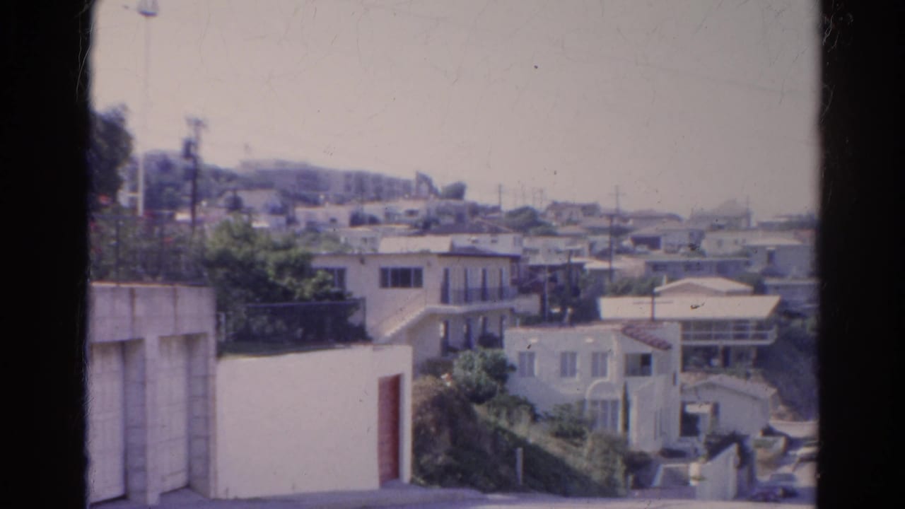 Residential hillside with pale houses, trees, power lines, and hazy sky.