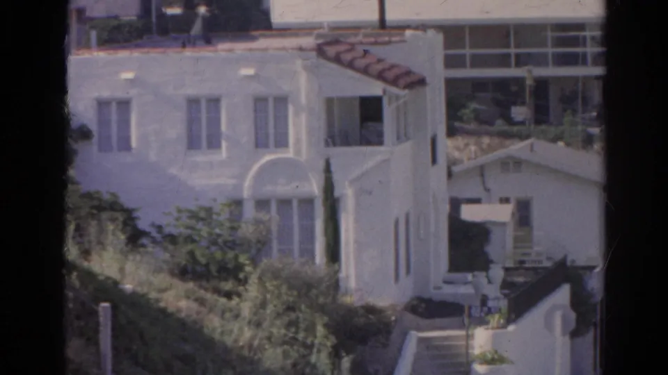 Closer 1966 Silver Lake view of a white stucco hillside house, shrubs, steps, neighboring roofs, and a street edge.