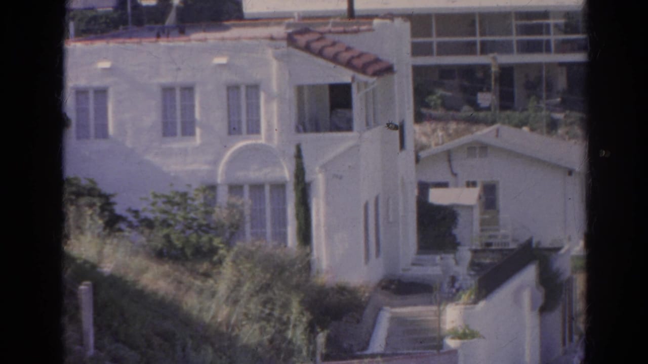 White stucco hillside house with arched window, greenery, and neighboring buildings.