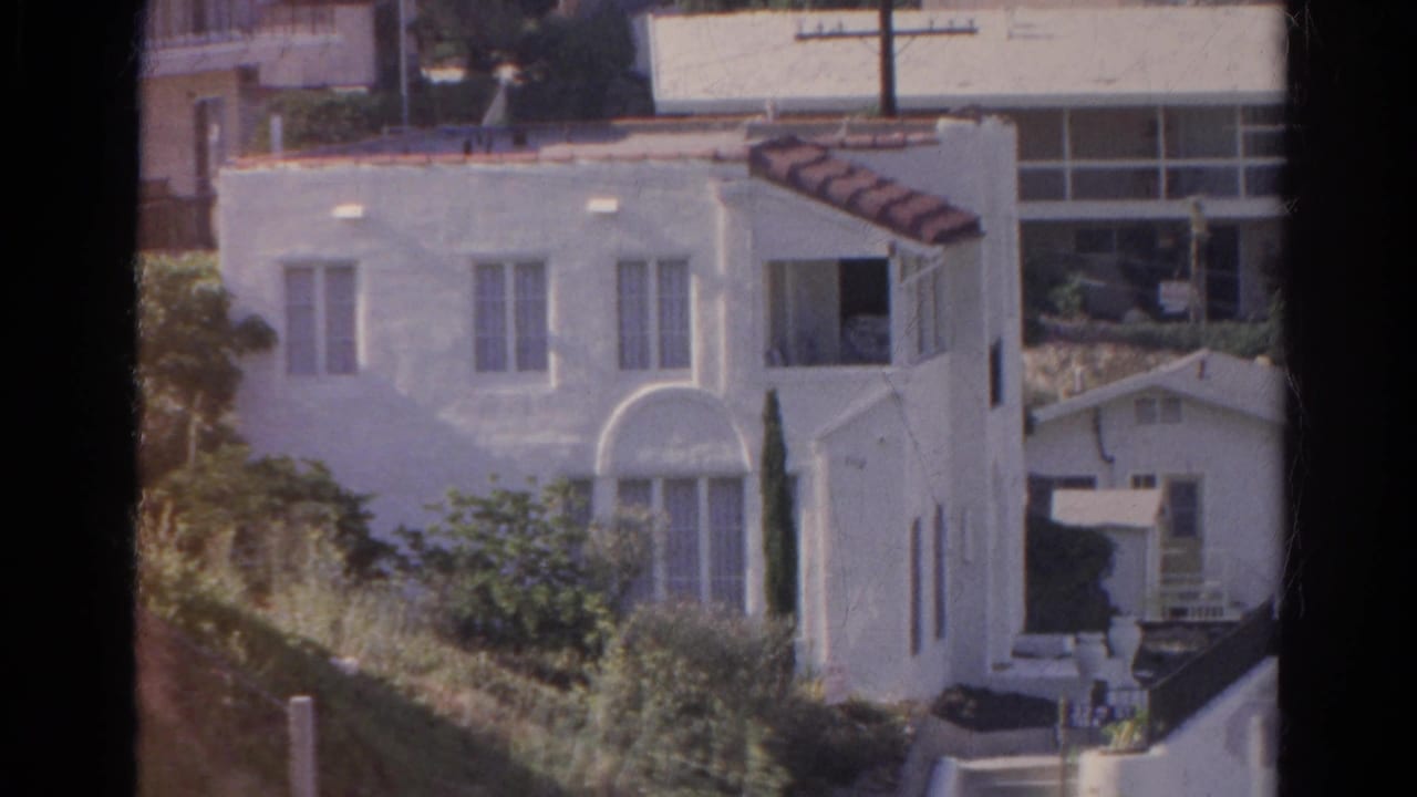 White hillside residence with arched window, tile roof edge, shrubs, and nearby homes.