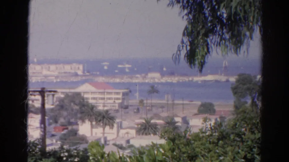 1966 8mm frame showing a sun-faded harbor overlook with foreground greenery, palms, rooftops, water, and distant white structures.