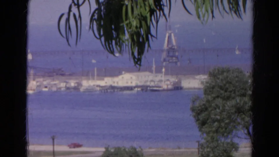 1966 8mm harbor overlook with a waterfront facility, moored boats, blue water, foreground foliage, and a small red car below.
