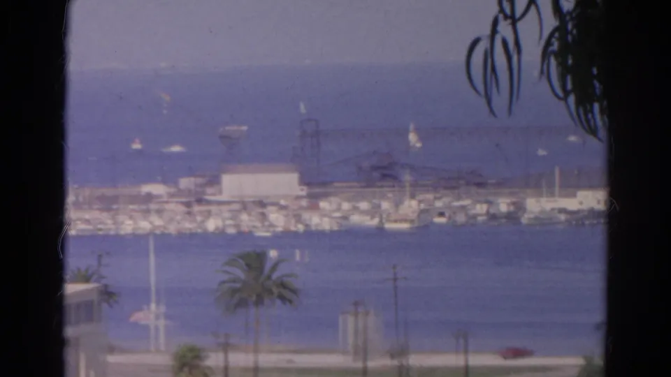 1966 8mm-style elevated California harbor view with blue water, sailboats, palm trees, and low waterfront buildings.