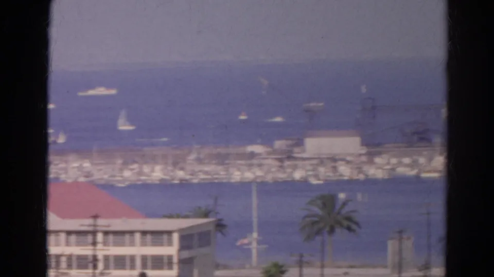 Elevated 1966 California waterfront view through a dark foreground divider, showing marina boats, blue water, and a red-roofed building.