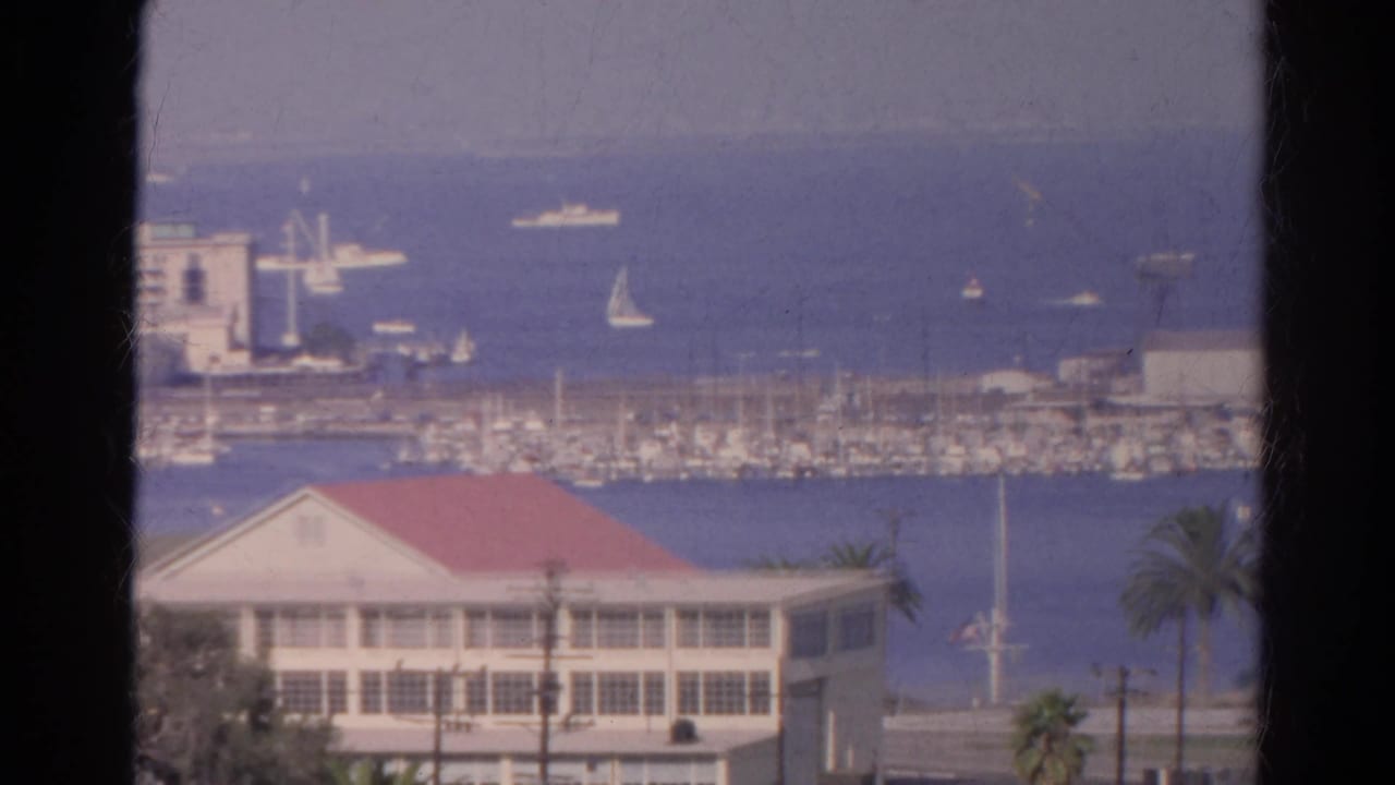 1966 8mm harbor scene with a red-roofed waterfront building, marina masts, sailboats, and palm trees in a coastal California view.