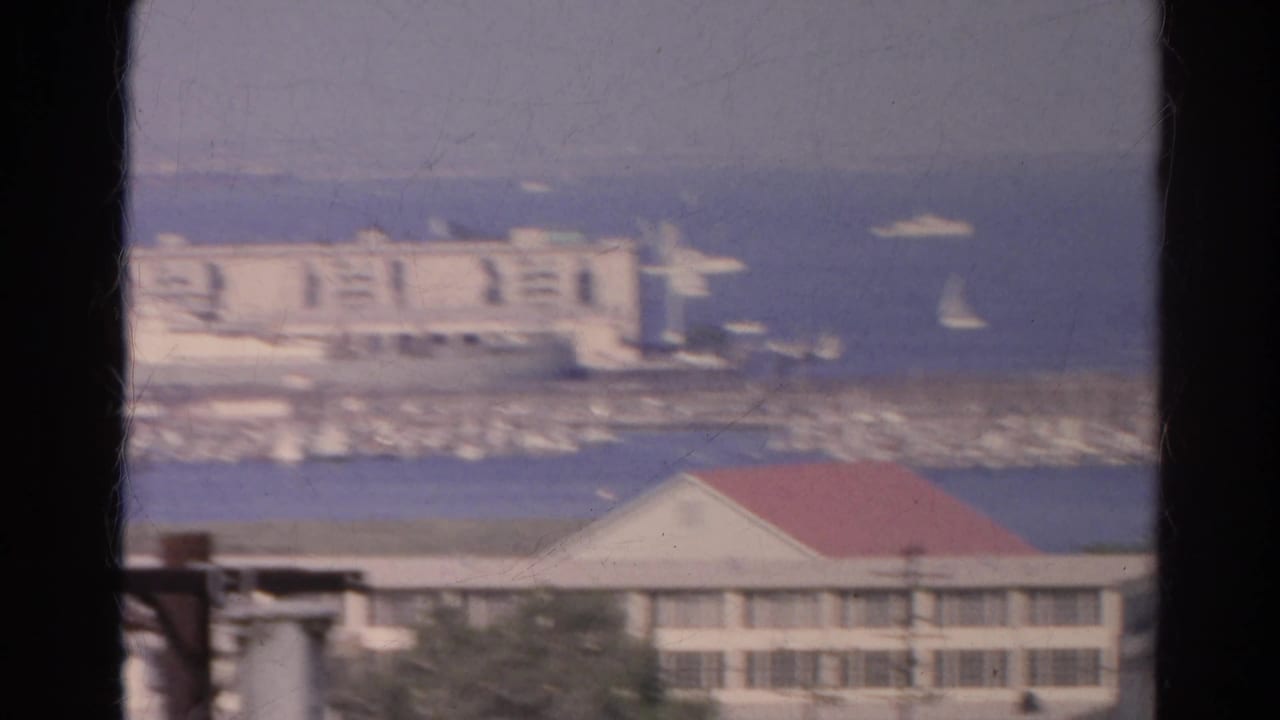 Soft 1966 8mm frame of a California harbor with a white waterfront structure, breakwater, sailboats, and a red-roofed building below.