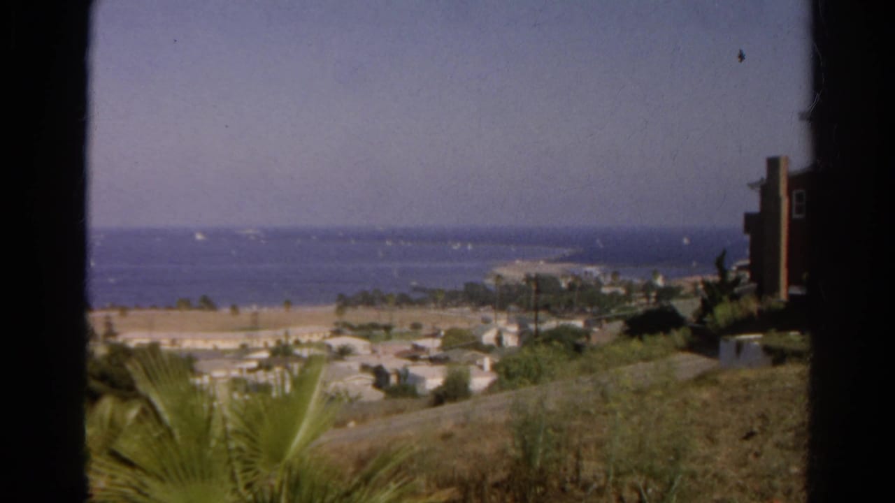 1966 California 8mm still showing a hillside view toward the ocean, with palms, low buildings, and small boats offshore.
