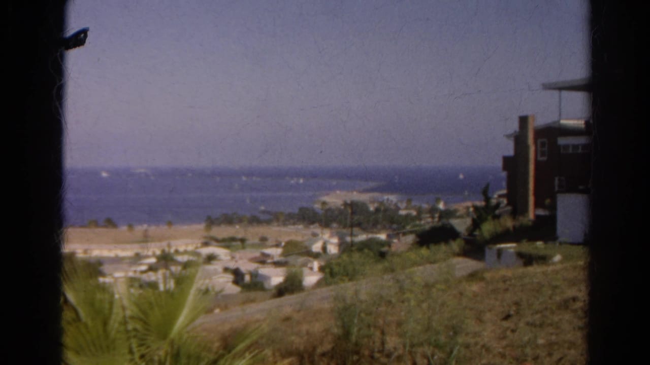 1966 California home-movie frame of a distant shoreline overlook, with dry hillside, palms, houses, and boats on the water.