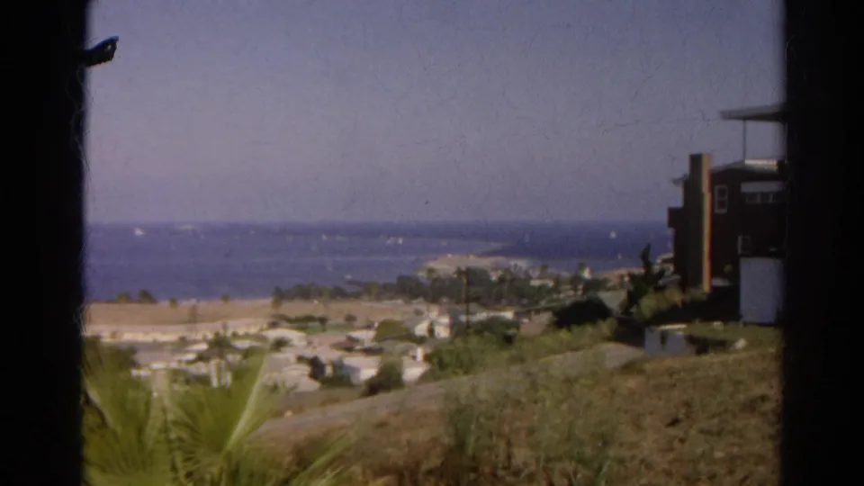 1966 California home-movie frame of a distant shoreline overlook, with dry hillside, palms, houses, and boats on the water.
