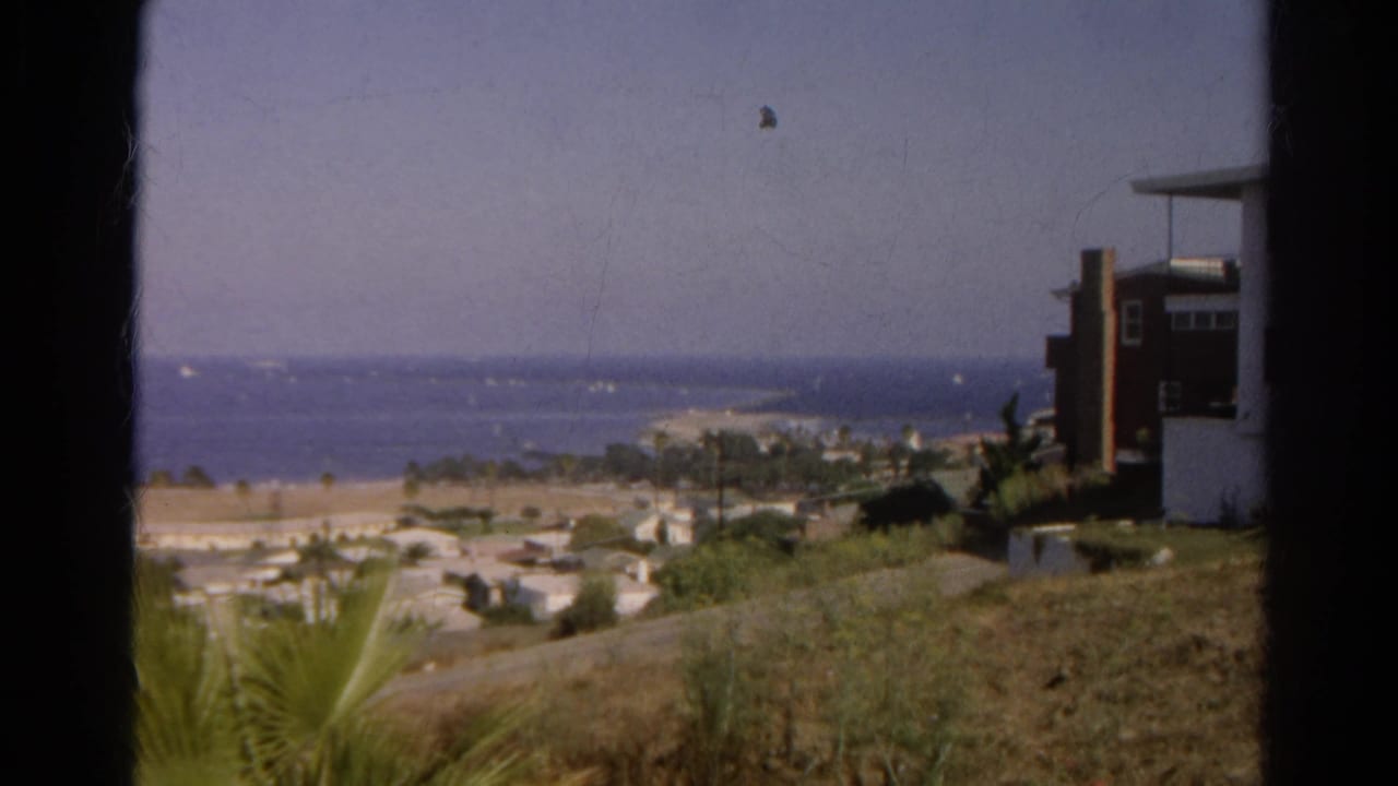 Elevated coastal view with beach houses, palms, ocean, sailboats, and dark frame edges.
