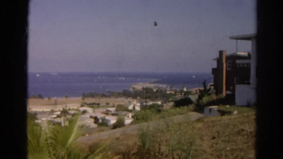 1966 8mm view identified as California, showing a coastal neighborhood, palms, beach, blue water, sailboats, and dark window-like borders.
