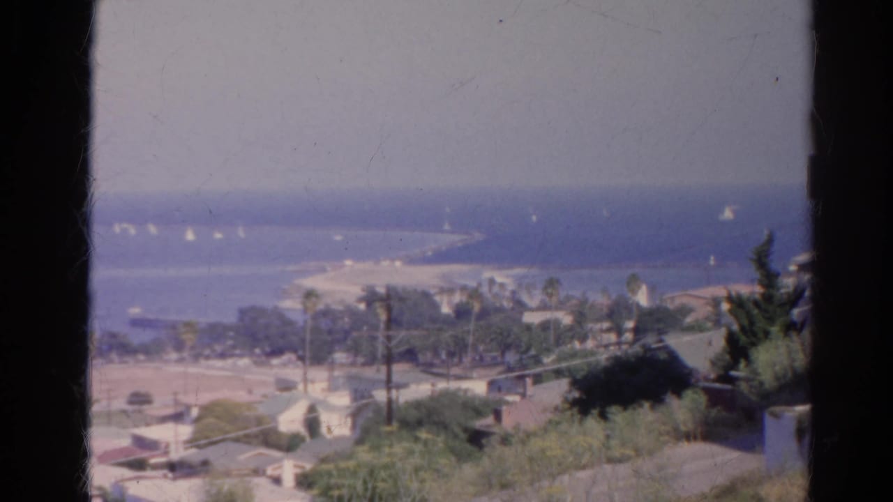 Soft-focus 1966 home-movie frame of a California shoreline, with residential rooftops, palms, beach sand, ocean, and scattered sailboats.