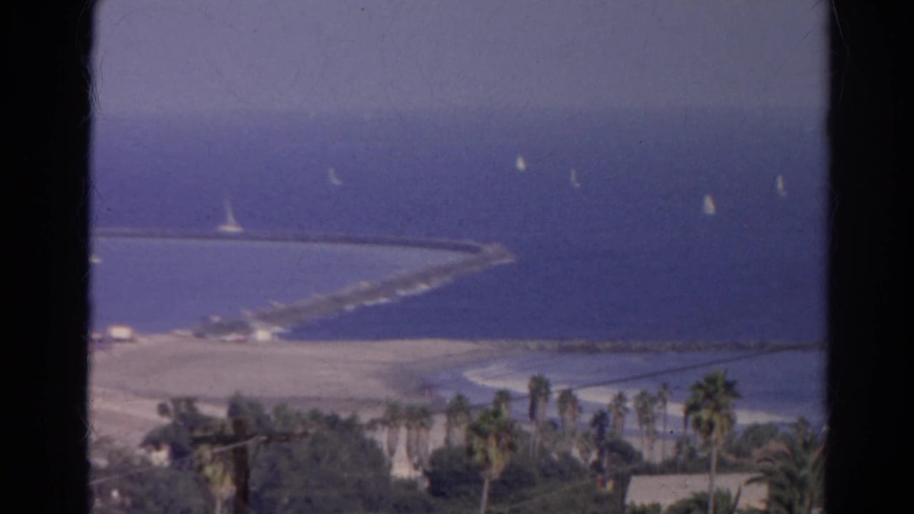 1966 8mm frame identified as a California coastal overlook, with sailboats beyond a breakwater and palms along the shore.