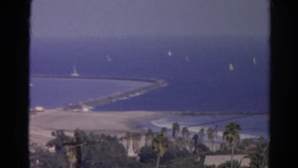 1966 8mm frame identified as a California coastal overlook, with sailboats beyond a breakwater and palms along the shore.