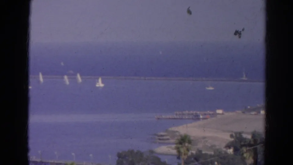 Vintage 8mm coastal frame from 1966 showing sailboats on blue water near a shoreline pier area, with palm trees and visible film wear.