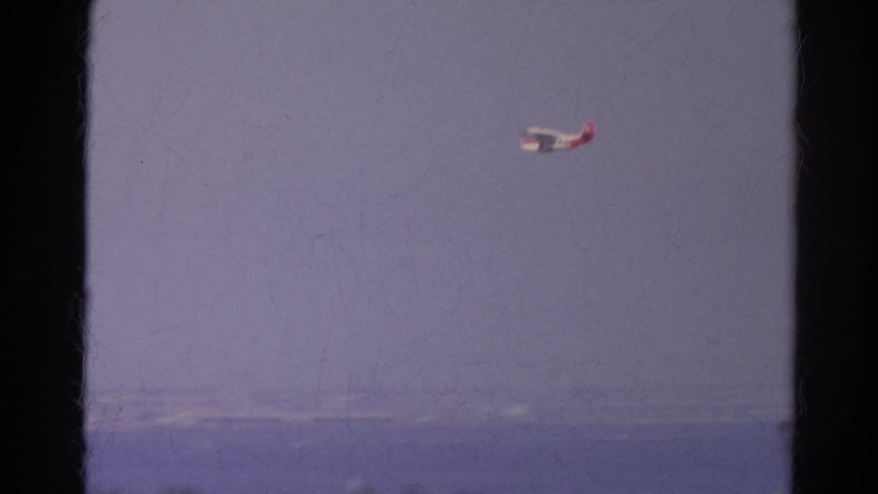 Small red and white airplane crossing hazy sky beyond dark window frames.
