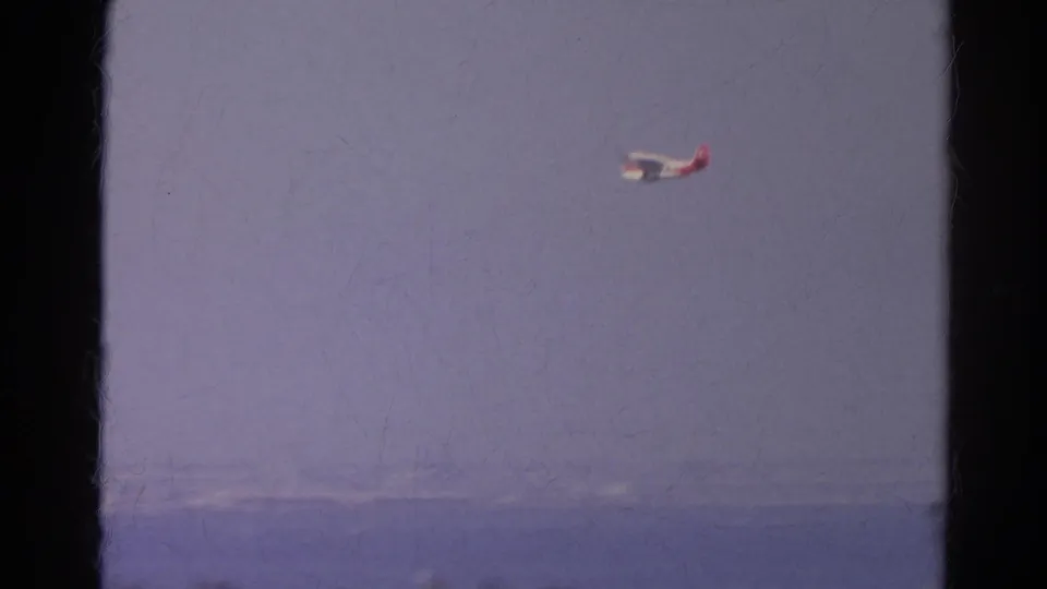 1966 8mm view of a small red and white airplane flying over a hazy California city and body of water, seen through window framing.