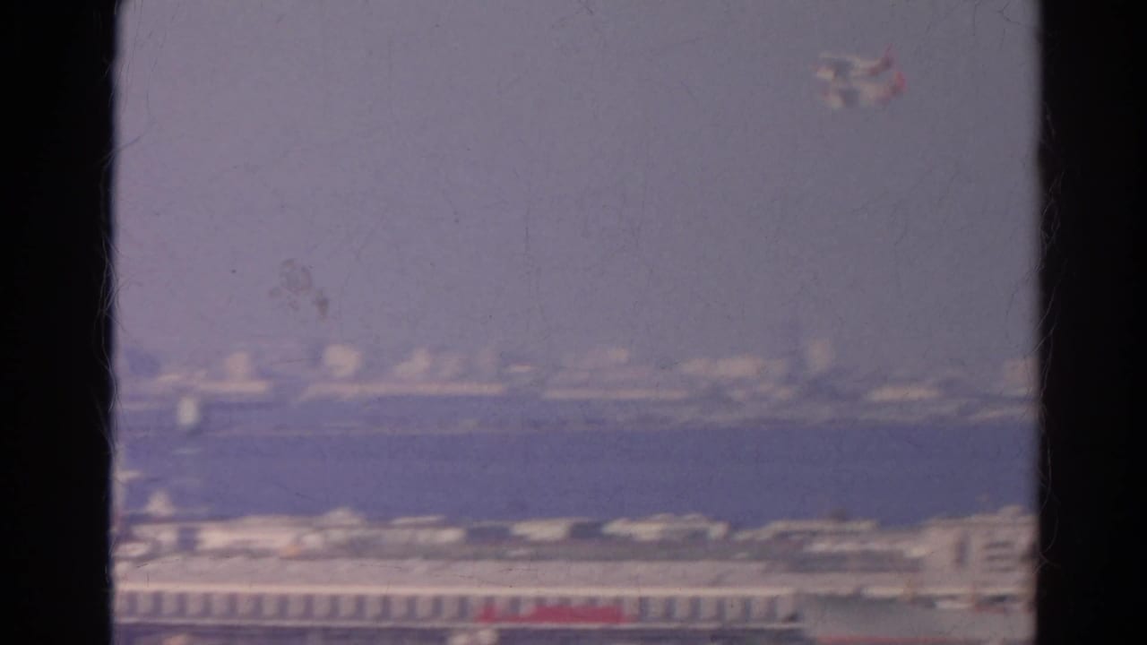 Small airplane appears high above distant waterfront buildings in a hazy 1966 California home-movie view through a window.