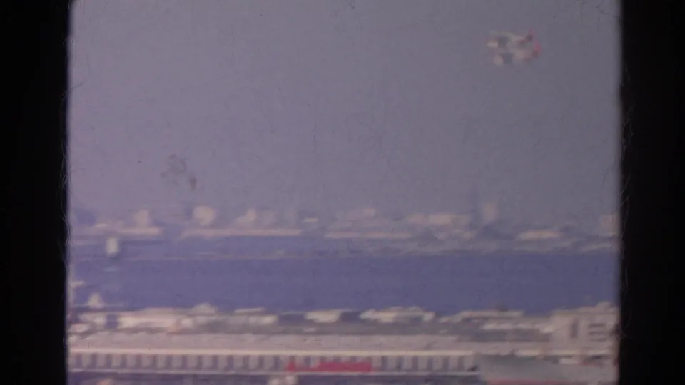 Small airplane appears high above distant waterfront buildings in a hazy 1966 California home-movie view through a window.
