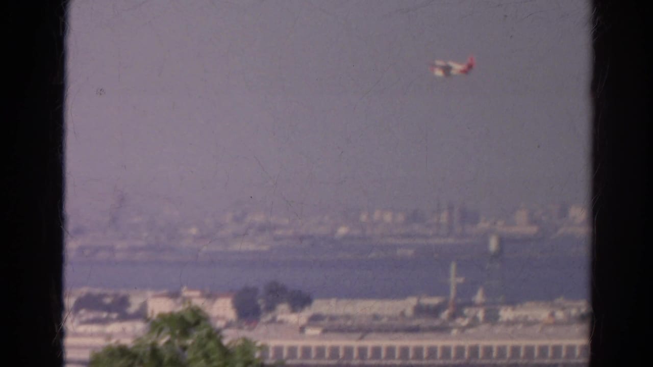 Red and white airplane crosses a hazy waterfront city view in 1966 8mm footage, with window framing and tree tops in the foreground.