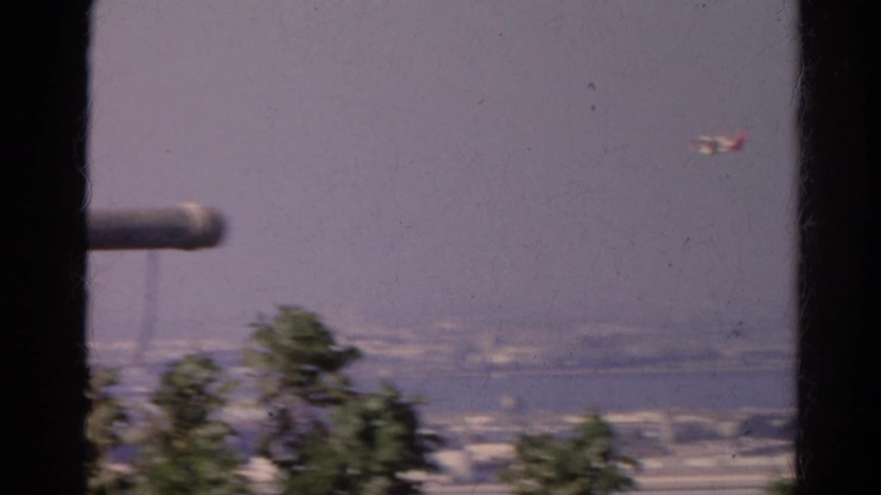1966 8mm frame shows a small airplane to the right of a window-framed waterfront city view, with blurred trees below.