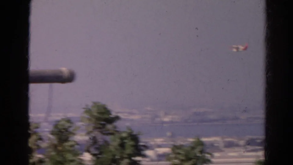 1966 8mm frame shows a small airplane to the right of a window-framed waterfront city view, with blurred trees below.