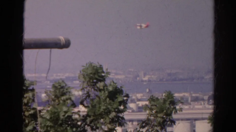 1966 California 8mm frame showing a small plane over a hazy shoreline with trees, distant water, and dark foreground framing.