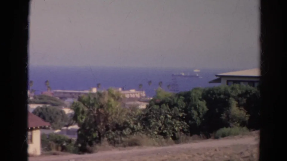1966 California 8mm view of a hazy ocean horizon with ships offshore, hillside vegetation, rooftops, and dark foreground framing.