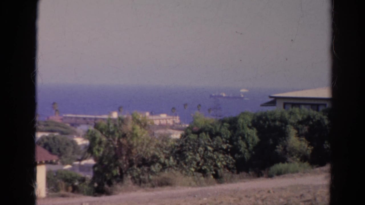 1966 California coastal frame showing distant ships on the water, a hazy sky, trees, rooftops, and a dark foreground divider.