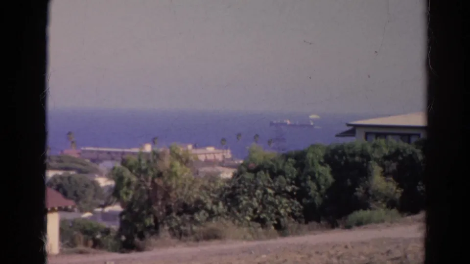 1966 California coastal frame showing distant ships on the water, a hazy sky, trees, rooftops, and a dark foreground divider.