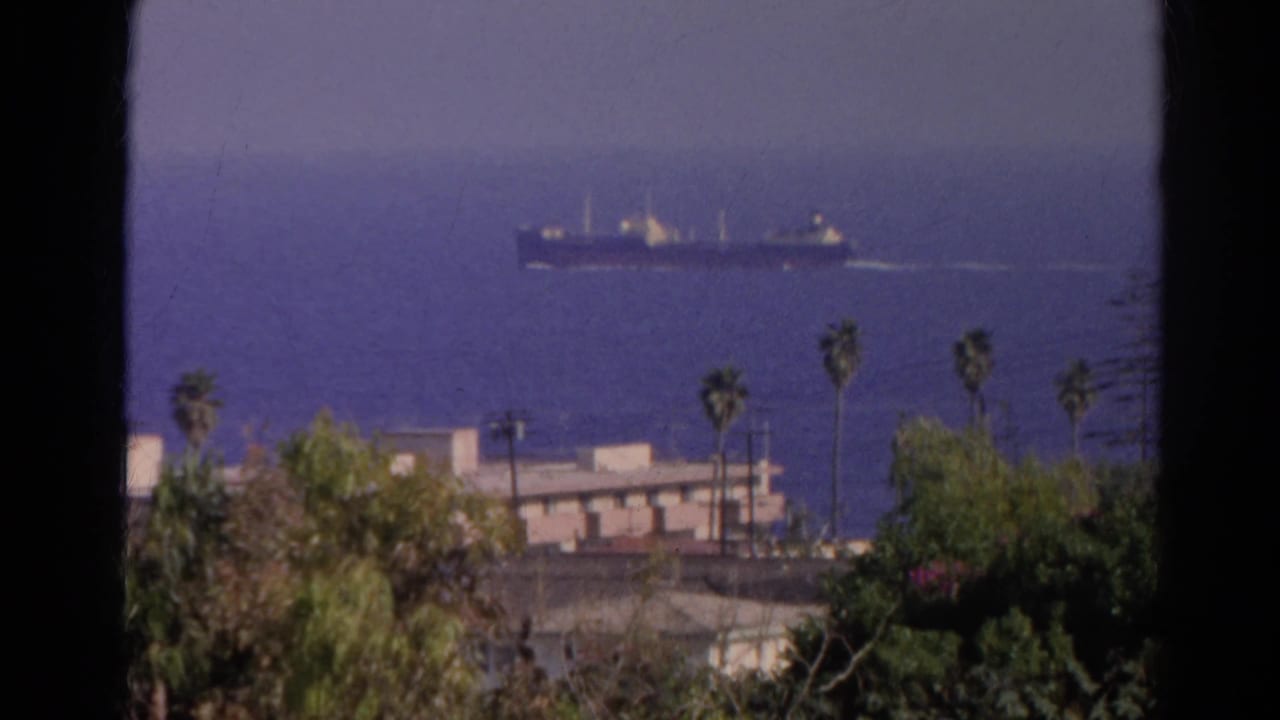 1966 8mm coastal shot with a large cargo ship offshore beyond palm trees, rooftops, and hillside vegetation.