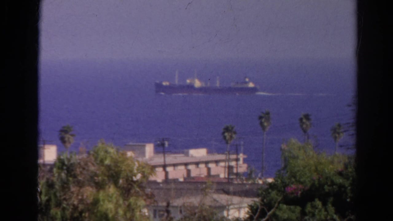 1966 California home-movie frame showing a cargo ship offshore with palms and rooftops below the horizon.