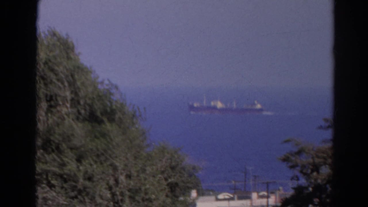 1966 8mm view of a distant cargo ship on blue water, framed by hillside trees, faint shoreline detail, and visible film gate edges.