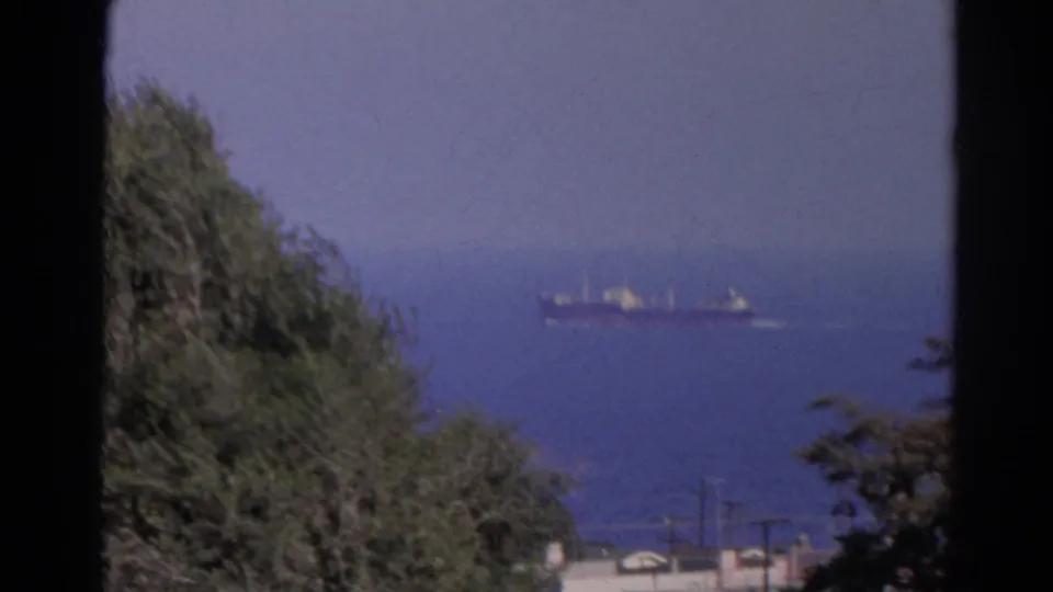 1966 8mm view of a distant cargo ship on blue water, framed by hillside trees, faint shoreline detail, and visible film gate edges.