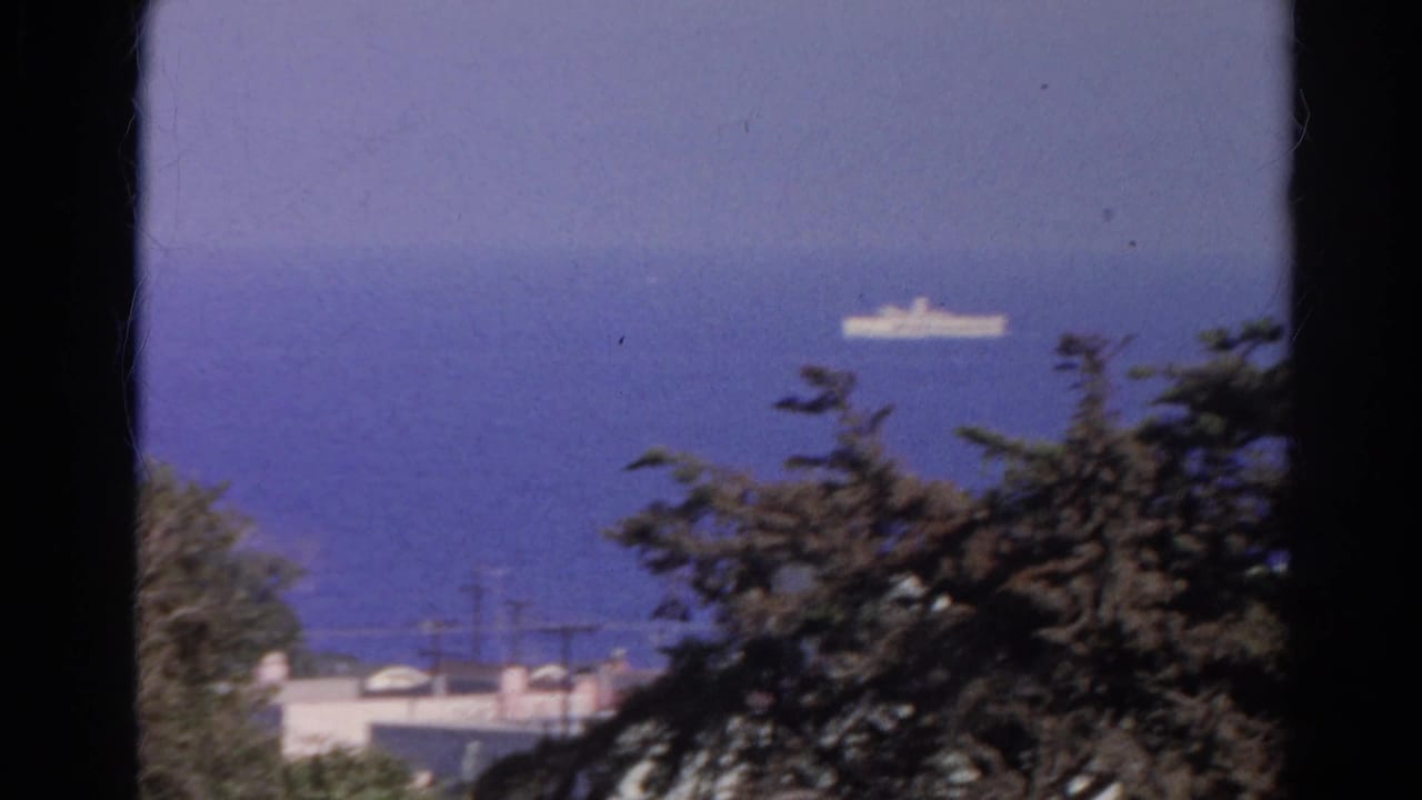 Distant white ship on blue water in 1966 8mm footage, viewed past trees, utility lines, and pale rooftops.