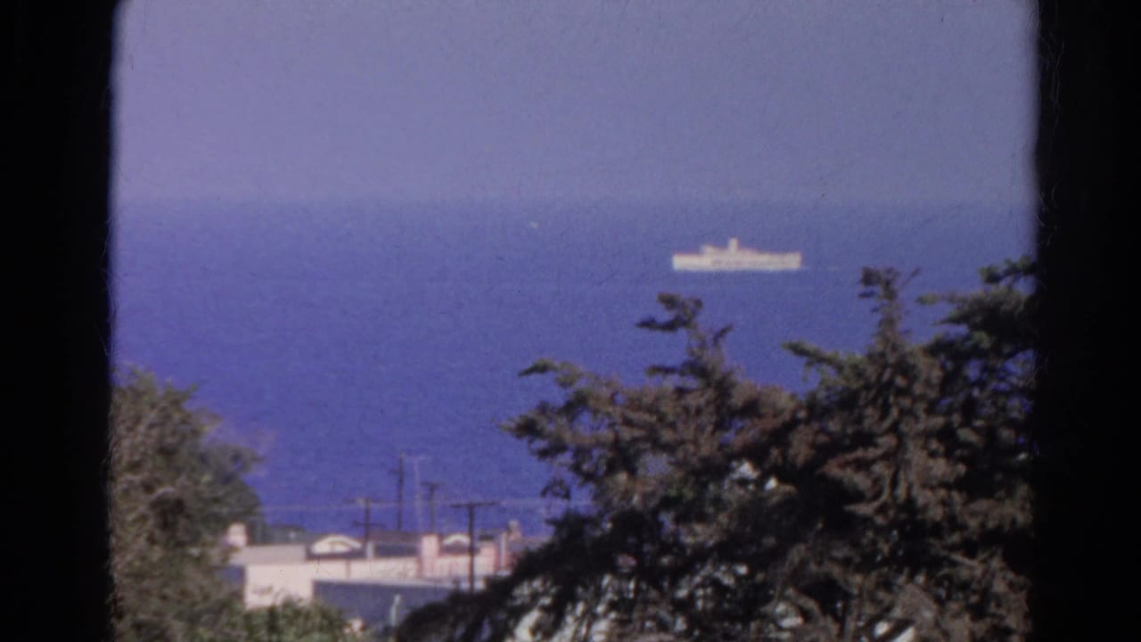 1966 8mm frame of a white ship offshore, with dark trees, blue water, and distant rooftops in the foreground.
