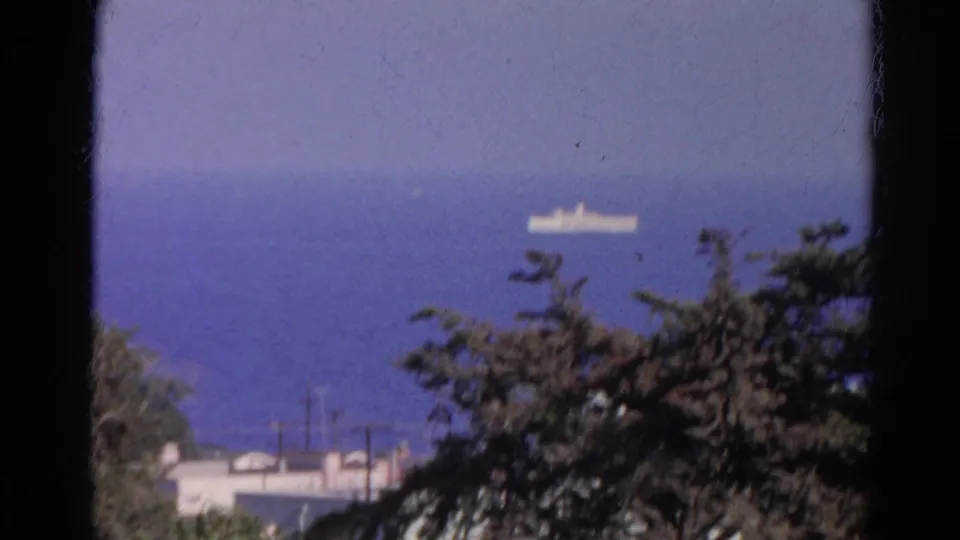1966 Silver Lake home-movie frame with a distant white ship on blue water, partially framed by trees and low residential rooftops.