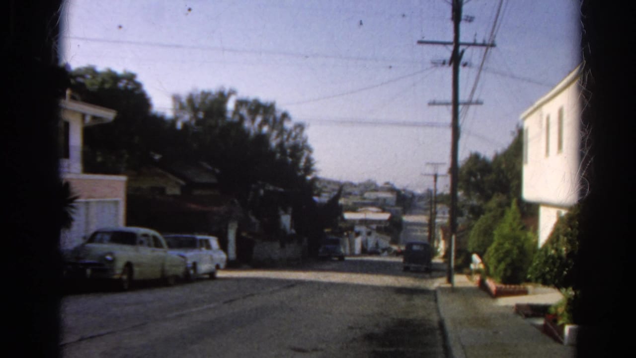 Quiet sloping residential street with parked cars, houses, utility poles, and distant hillside homes.