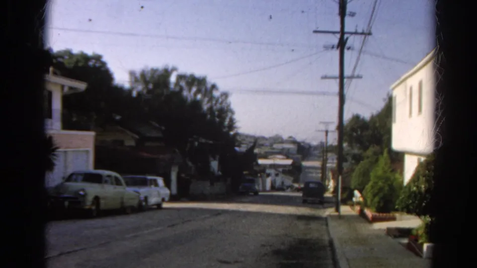 1966 Silver Lake home-movie view of a sunlit residential street with parked cars, utility poles, houses, and hillside homes in the distance.