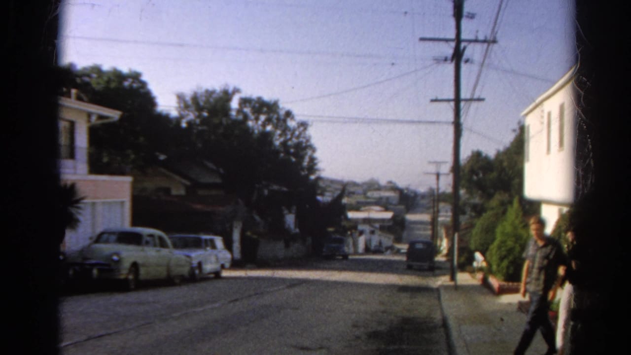 Residential street with parked cars and a person entering at the right edge near a white building.