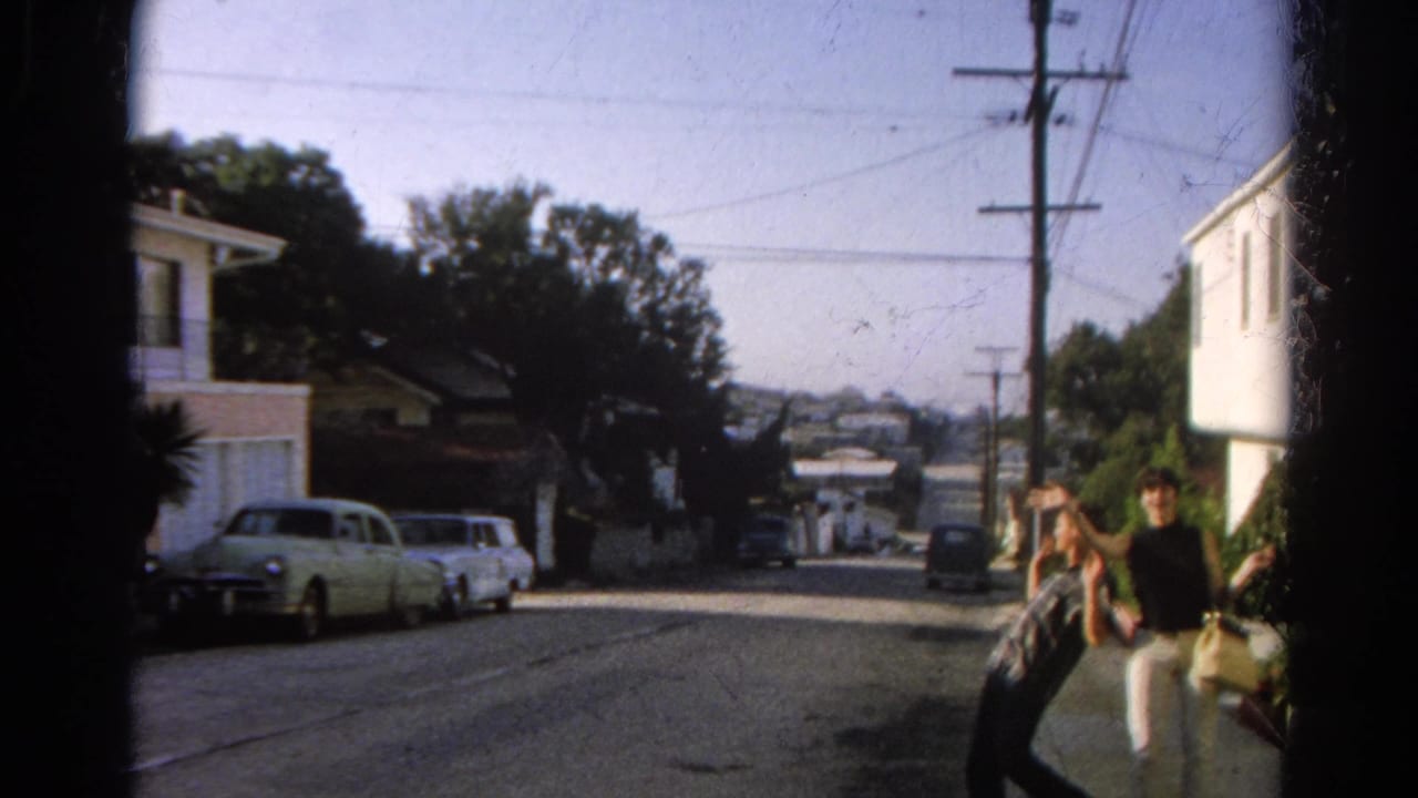 Two pedestrians pass through a 1966 Silver Lake residential street view with parked cars, houses, utility poles, and bright afternoon light.