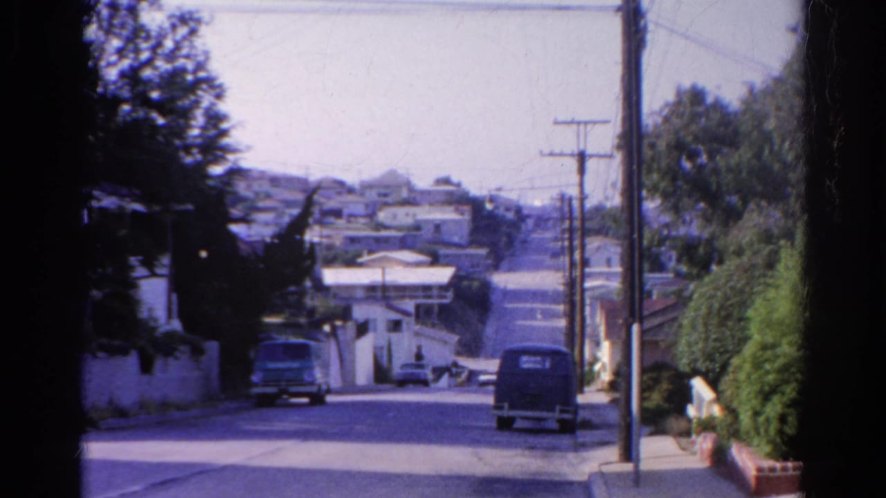 End frame looking down a Silver Lake hillside street with parked vehicles, utility poles, hedges, and distant residential buildings.