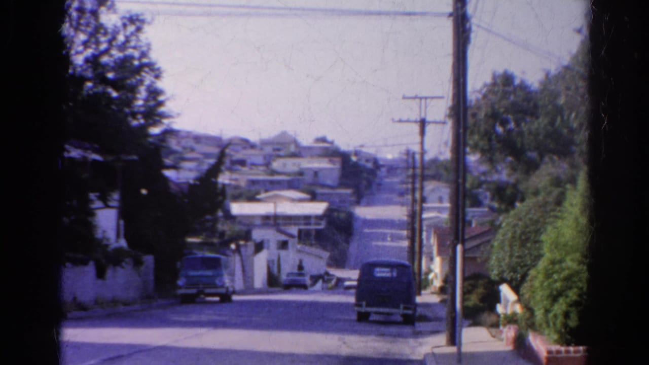 Sloped residential street with parked cars, utility poles, trees, and hillside houses.