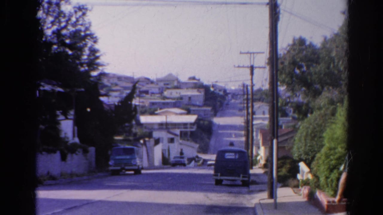 Residential hillside road with parked vehicles, utility poles, trees, and houses.