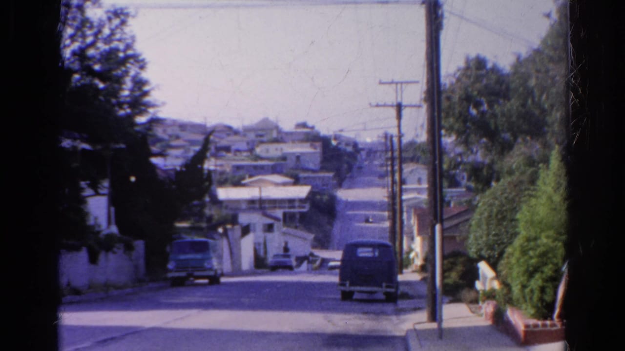 1966 Silver Lake home-movie frame of a sloping residential street with parked cars, stucco walls, utility poles, and distant hillside houses.